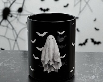 May include: Black ceramic mug featuring a white ghost design with bats. The ghost is draped in a sheet with two eye holes. White bats are scattered around the ghost on the black mug. Halloween themed.
