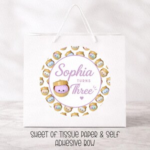 May include: White gift bag with a circular design featuring the name "Sophia" and the words "Turns Three." The design includes a cartoon illustration of a small container with a cute face. The bag also has the text "SHEET OF TISSUE PAPER & SELF ADHESIVE BOW."