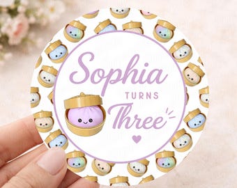 May include: A round sticker with a white background and a purple border. The sticker features the name "Sophia" and the phrase "Turns Three" in purple script. The design includes cartoon dumplings in bamboo steamers.