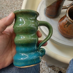 May include: A handmade ceramic mug with a green and blue glaze. The mug has a unique, rounded shape and a handle.