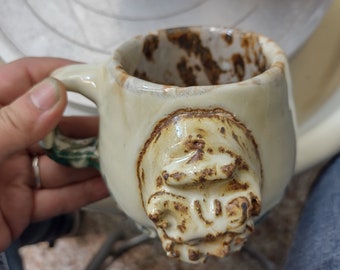 May include: A white ceramic mug with a brown glaze and a sculpted face on the handle. The mug is sitting on a pottery wheel.