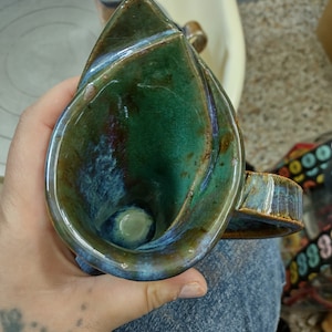 May include: A green and blue ceramic mug with a unique, swirling design. The mug has a handle and a wide, open mouth.