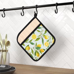 May include: A square pot holder with a black border and a light beige top section. The main section features a pattern of lemons, green leaves, and white flowers on a light blue background. It hangs from a black hook.