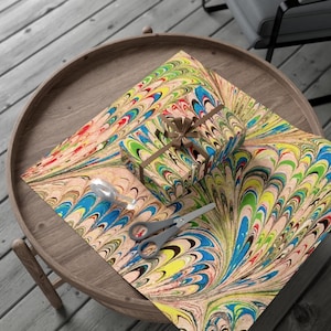 May include: A wrapped gift box and wrapping paper with a colorful, swirled pattern in shades of blue, green, yellow, and red. The gift is tied with a brown ribbon. A pair of scissors and a roll of tape are on the paper, all on a wooden table.