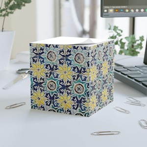 May include: A square notepad holder with a repeating pattern of blue, yellow, and teal floral designs. The notepad holder sits on a white surface with scattered silver paper clips and a computer keyboard in the background.