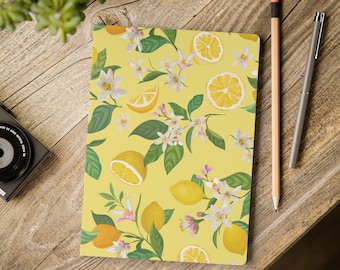 May include: A yellow notebook with a lemon and floral pattern. The design features whole and sliced lemons, white blossoms, and green leaves. A pencil and pen are next to the notebook on a wooden surface.