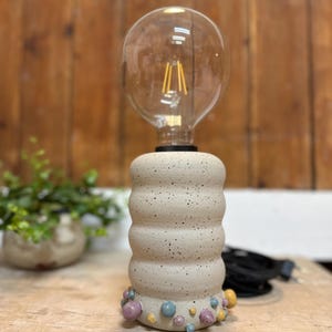 Könnte beinhalten: Eine Tischlampe aus Keramik mit einer großen, runden Glühbirne. Der Lampensockel ist beige mit einem strukturierten, gestapelten Design und farbenfrohen, abgerundeten Akzenten. Ein schwarzes Kabel ist sichtbar.