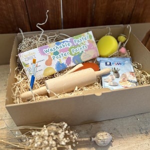 Op de afbeelding: Een bruine kartonnen doos gevuld met knutselspullen. De doos bevat een deegroller, twee kwasten, een pakket pastelposterverf, een pakket polymer clay, een gele spons en een roze spons.