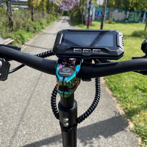 Puede incluir: Manillar negro de patinete eléctrico con pantalla digital y abrazadera colorida. La pantalla muestra información. El manillar es negro con un cable en espiral y una abrazadera con el texto "Carbonic V6".