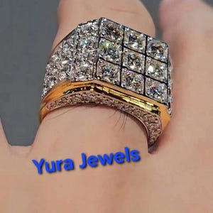 May include: A gold and silver ring with a large square top, encrusted with numerous small, sparkling diamonds. The ring has a gold band and is set with multiple rows of diamonds. The text "Yura Jewels" is visible.