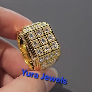 May include: A gold-colored ring with a square top, encrusted with multiple clear, square-cut gemstones. The ring has a band of smaller gemstones around the sides. The text "Yura Jewels" is visible in blue. Jewelry.