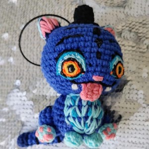 Puede incluir: Una criatura azul tejida a ganchillo con forma de gato, con ojos naranjas y amarillos, una lengua rosa y un pequeño sombrero de copa negro. El peluche hecho a mano tiene un diseño azul claro y rosa en las patas y el pecho.