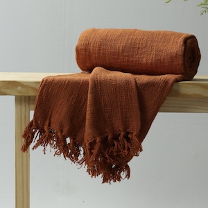 May include: A rust-coloured throw blanket with fringe on a wooden bench. The blanket is folded and rolled up on the bench.