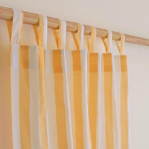 May include: Yellow and white striped curtains hanging from a wooden rod. The curtains have a simple design with vertical stripes and fabric loops for hanging. The color scheme is bright and cheerful, suitable for a variety of interior styles.