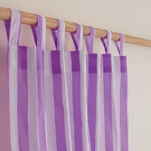 May include: Striped curtain panels in shades of purple and white hang from a wooden rod. The curtains feature vertical stripes and fabric loops for hanging. The color scheme is a soft, inviting combination, suitable for a bedroom or living room.