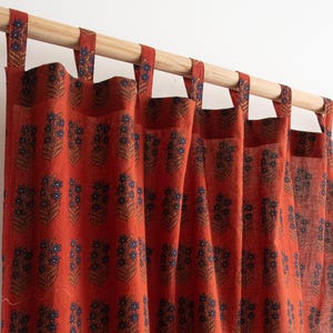 May include: Terracotta-coloured curtains featuring a repeating floral pattern in navy and brown. The curtains hang from a wooden pole with matching fabric loops. The floral design is a stylized depiction of a cluster of flowers.