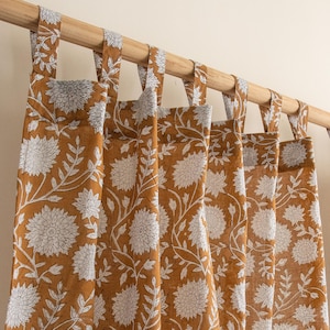 Puede incluir: Cortinas color mostaza con estampados florales blancos. Las cortinas cuelgan de una barra de madera con presillas de tela a juego. El diseño floral se repite en los paneles.