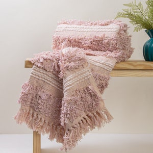 Può includere: Una coperta in tessuto rosa e bianco con frange. La coperta è appoggiata su una panca di legno.