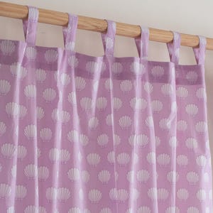 Puede incluir: Cortinas de color lila con un patrón repetido de conchas blancas. Las cortinas cuelgan de una barra de madera con lazos de tela lila a juego. La tela tiene un aspecto suave y fluido.
