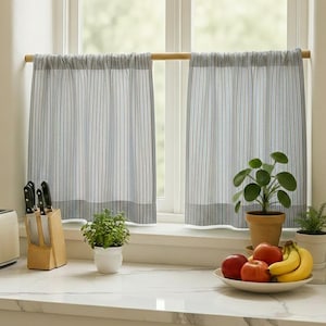 May include: A kitchen window with blue and white striped curtains. A toaster, knife block, and a fruit bowl with apples and bananas are on the counter. Small potted plants add a touch of greenery to the scene.