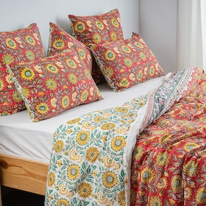 Op de afbeelding: Een wit bed met een rood en geel bloemenpatroon dekbedovertrek en bijpassende kussenslopen. Het bed is gemaakt met een wit laken en heeft een houten frame.