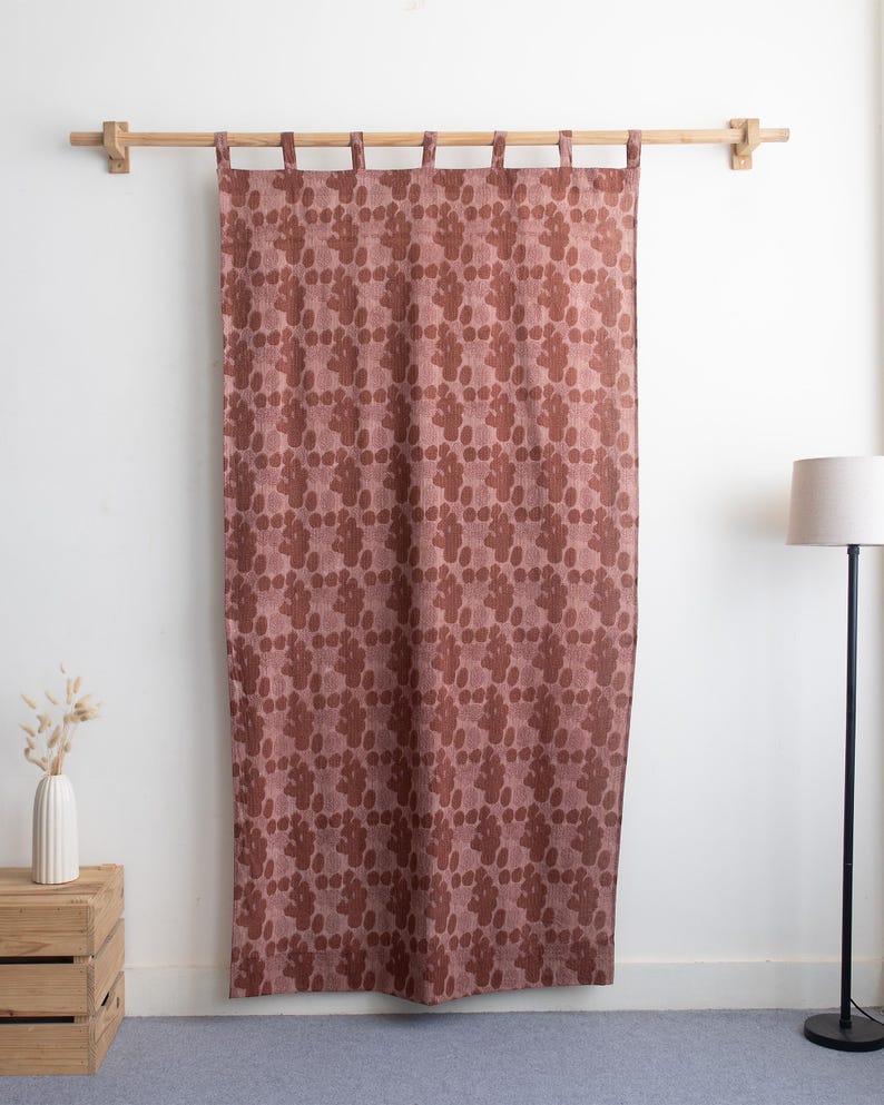 Puede incluir: Una cortina rosa polvoriento con un patr&oacute;n repetitivo de formas org&aacute;nicas marr&oacute;n oscuro. La cortina cuelga de una barra de madera natural con soportes de madera a juego. La cortina est&aacute; hecha de una tela texturizada.
