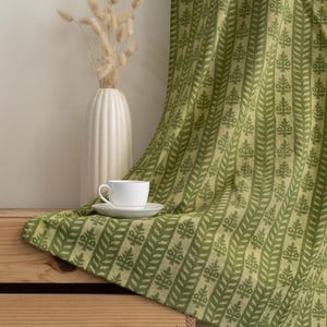 May include: A white teacup and saucer sit on a green patterned fabric draped over a wooden crate. The fabric features a repeating leaf design in varying shades of green. A white vase with dried flowers is in the background.