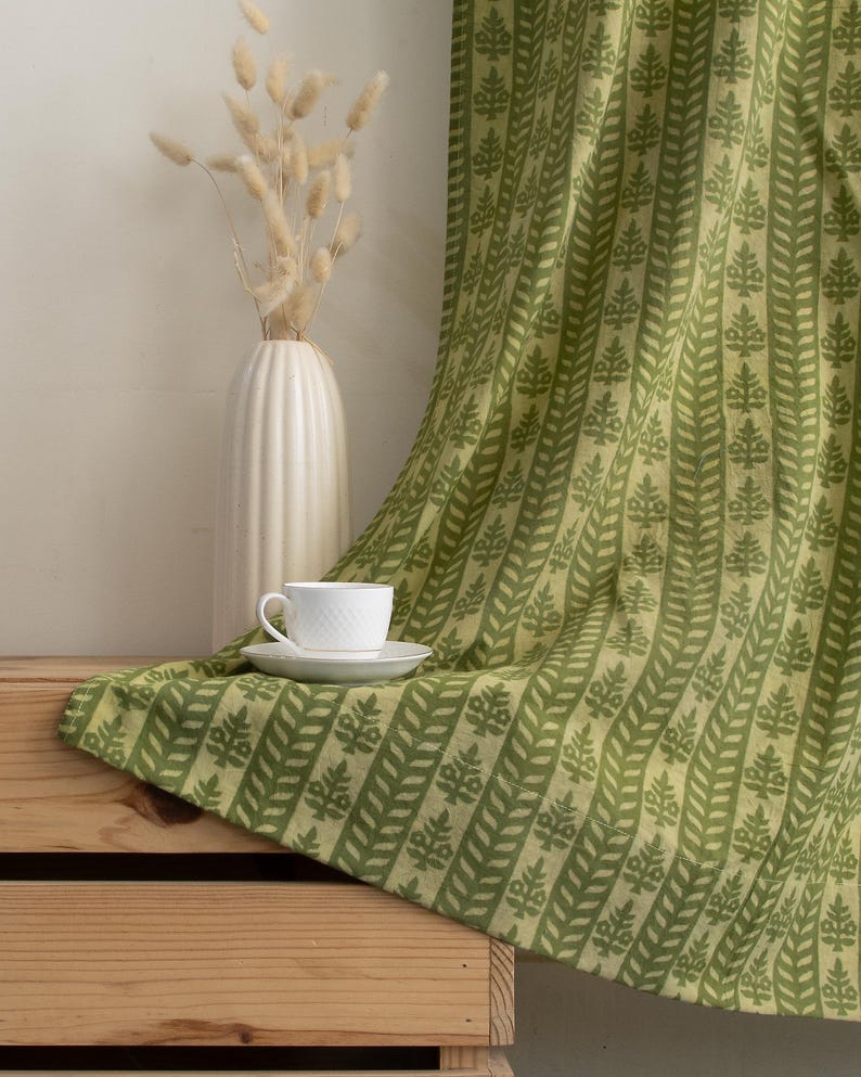 May include: A white teacup and saucer sit on a green patterned fabric draped over a wooden crate. The fabric features a repeating leaf design in varying shades of green. A white vase with dried flowers is in the background.