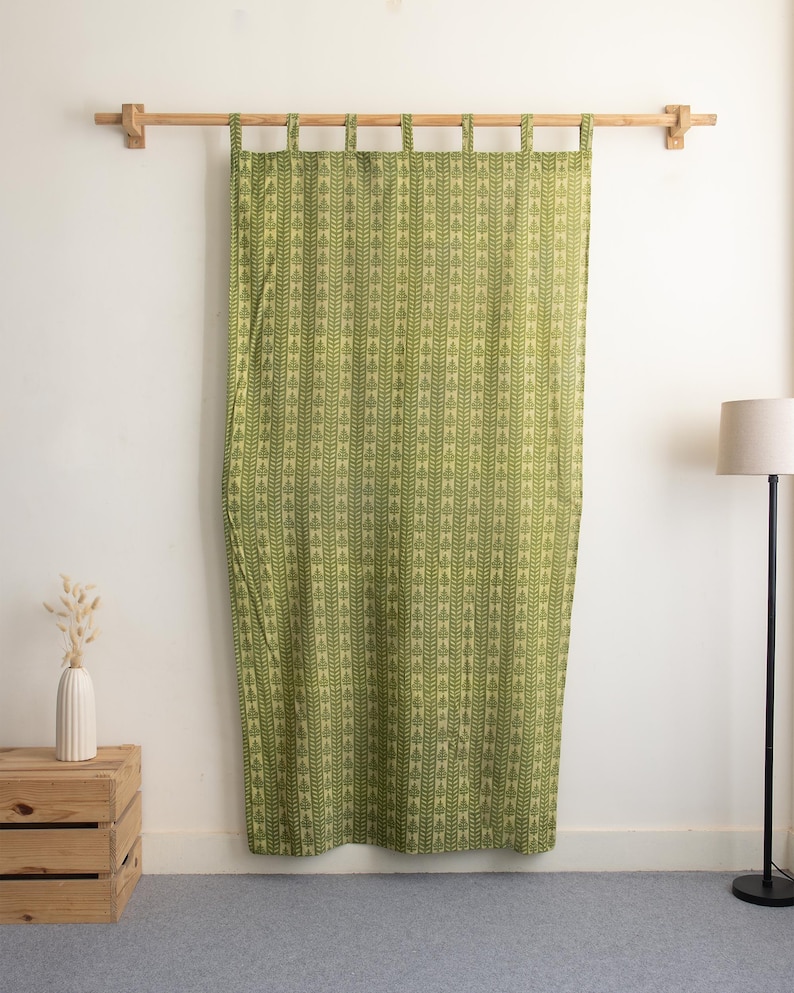 May include: Green curtain with a repeating leaf pattern, hanging from a wooden rod. The curtain is a light green color with darker green leaf designs. The curtain is hanging in front of a white wall.