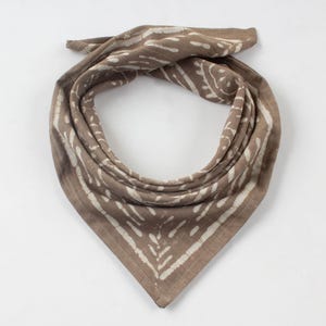 May include: A brown bandana with a white, patterned design. The square scarf is folded in half diagonally and tied at the top. The fabric has a soft, textured appearance, and the white pattern is evenly distributed across the surface.
