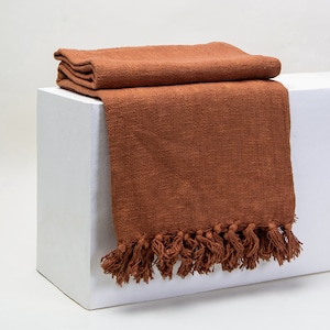 May include: A rust-coloured throw blanket with a fringe border. The blanket is folded and displayed on a white surface.