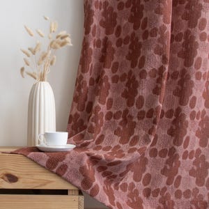 Puede incluir: Una cortina drapeada con un estampado floral en tonos rosa empolvado y marr&oacute;n es el foco principal. Un motivo floral se repite en la tela. Una taza y un platillo blancos descansan sobre una caja de madera, junto a un jarr&oacute;n blanco con flores secas. La escena evoca calidez.
