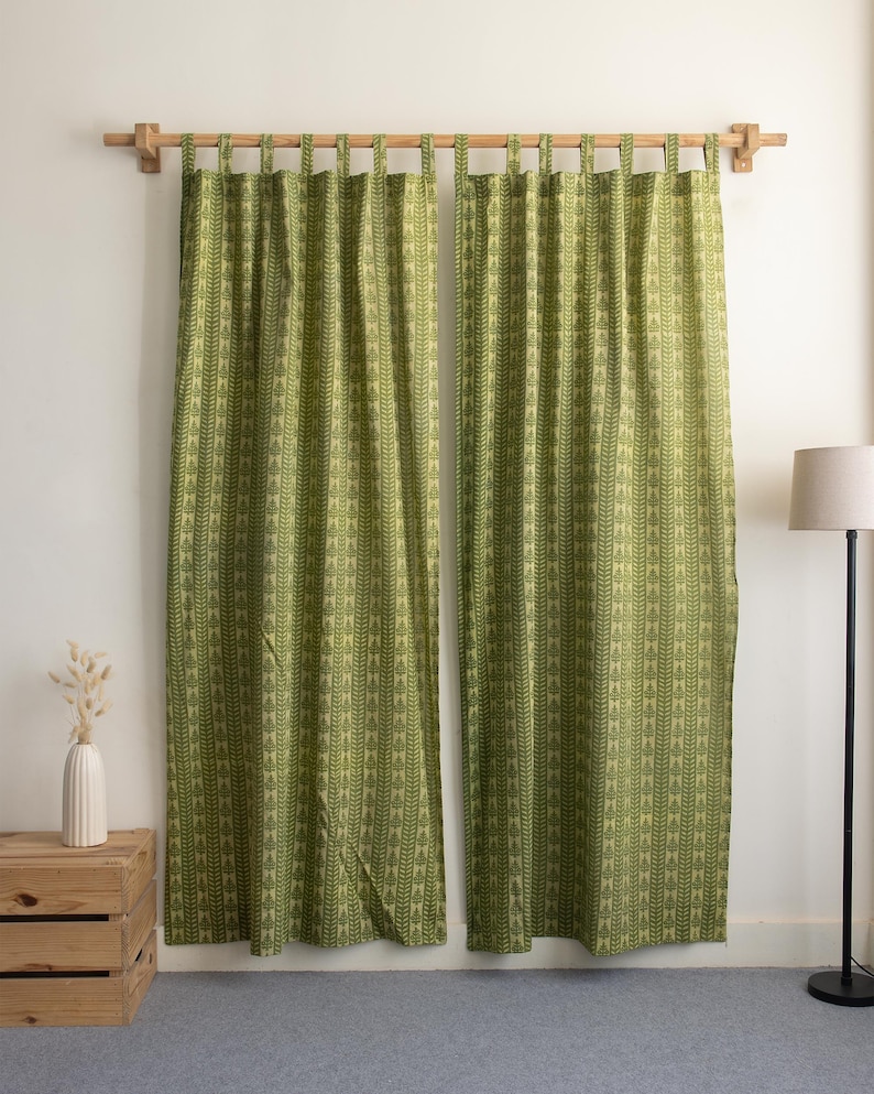 May include: Two green curtains with a repeating leaf pattern hang from a wooden rod. The curtains are a light green color with a darker green leaf design. The curtains are hanging in front of a white wall.