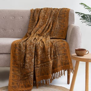 Peut inclure: Plaid de couleur moutarde avec un motif floral et géométrique drapé sur un canapé gris. La couverture a un bord à franges. Une petite table d'appoint en bois contient une tasse en céramique marron.