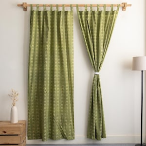 May include: Two green patterned curtains hanging from a wooden rod. The curtains have a repeating leaf design. A white cord holds one curtain open. A wooden crate and a floor lamp are in the background.