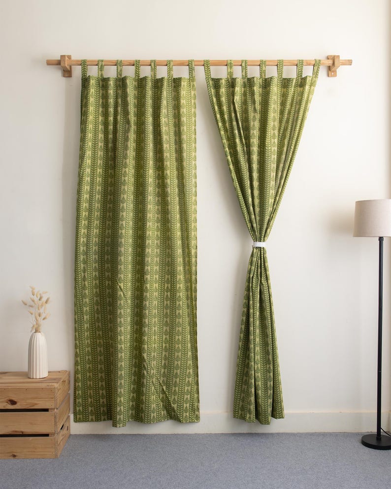 May include: Two green patterned curtains hanging from a wooden rod. The curtains have a repeating leaf design. A white cord holds one curtain open. A wooden crate and a floor lamp are in the background.