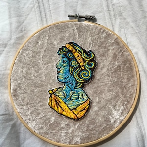 Könnte beinhalten: Gesticktes Porträt einer Person mit blauen und gelben wirbelnden Mustern in einem hölzernen Stickrahmen auf weißem Hintergrund.