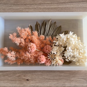 May include: A white rectangular tray with a dried floral arrangement. The arrangement includes peach-colored flowers, white flowers, and dried palm leaves.