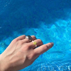 May include: A hand wearing two rings. The ring on the index finger is a silver band. The ring on the middle finger is a white beaded ring with a yellow smiley face charm.