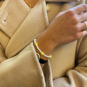 Könnte beinhalten: Ein weißes Perlenarmband mit gelben Smiley-Anhängern. Das Armband befindet sich am Handgelenk.