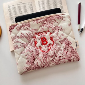 Könnte beinhalten: Cremefarbene, gesteppte Tasche mit rotem Toile-de-Jouy-Muster mit Figuren und Bäumen. Die Tasche hat ein rotes, gesticktes "B" in einem dekorativen Rahmen. Die Tasche ist offen und zeigt einen schwarzen Gegenstand im Inneren.