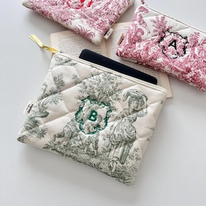 Könnte beinhalten: Gesteppte Täschchen mit Toile-de-Jouy-Muster in Creme und Burgunderrot. Ein Täschchen hat ein grün gesticktes "B", ein anderes ein schwarz gesticktes "A". Die Täschchen liegen auf einem offenen Buch.