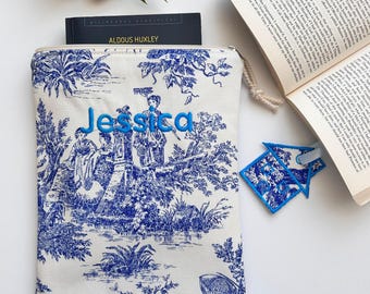 Può includere: Una custodia per libri bianca con un motivo toile blu e il nome "Jessica" ricamato su di essa. La custodia è appoggiata su una superficie bianca accanto a un libro e un segnalibro blu a forma di casa.