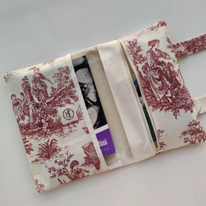 May include: A white fabric book clutch with a red and white floral pattern. The clutch has a zipper closure and a flap with a button closure. The clutch is open to reveal a purple book with the text "t&uuml;su" on the cover.