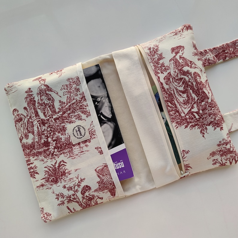 May include: A white fabric book clutch with a red and white floral pattern. The clutch has a zipper closure and a flap with a button closure. The clutch is open to reveal a purple book with the text "t&uuml;su" on the cover.