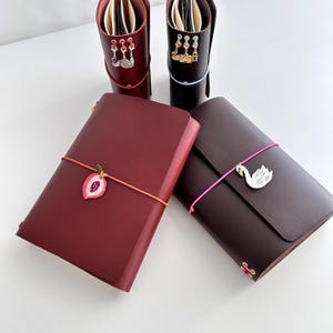 May include: Two dark red and two dark brown leather journals with elastic bands. The journals feature metal charms including a swan, a peach, and various small shapes attached to the elastic closures and the top edges of the journal covers.