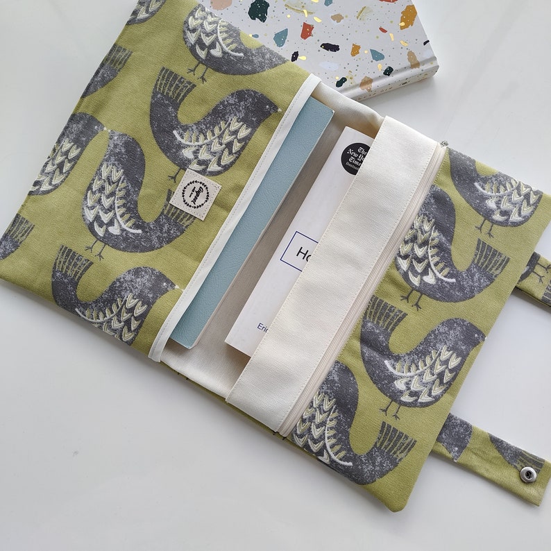 K&ouml;nnte beinhalten: Eine gr&uuml;ne Stofftasche mit Rei&szlig;verschluss und einem Vogelmuster. Die Tasche ist offen und enth&auml;lt zwei Notizb&uuml;cher. Die Notizb&uuml;cher sind wei&szlig; und blau.