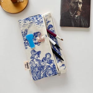 以下が含まれることがあります： 風景の中に人物を描いた、青と白のトワルドジュイ柄のキャンバス製ペンケース。 ペンケースは部分的に開いており、ペン、青いマーカー、ペーパークリップが見えます。肖像画のノートも表示されています。