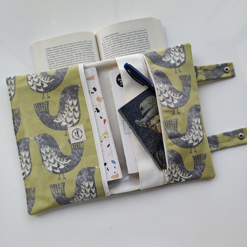 K&ouml;nnte beinhalten: Eine gr&uuml;ne Stofftasche f&uuml;r B&uuml;cher mit einem Vogelmuster. Die Tasche hat einen Rei&szlig;verschluss und ist ge&ouml;ffnet, um ein Buch und ein Notizbuch im Inneren zu zeigen.