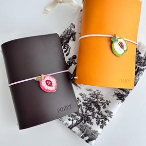 May include: Two leather journals, one dark brown with the name POPPY and one orange with the name DANA embossed on the front. Each journal is secured with an elastic band featuring a stitched fruit charm, a peach on the brown journal and an apple on the orange one. They rest on a white and black patterned fabric pouch.